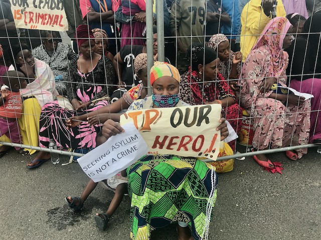 Photo of refugees with placards behind a fence